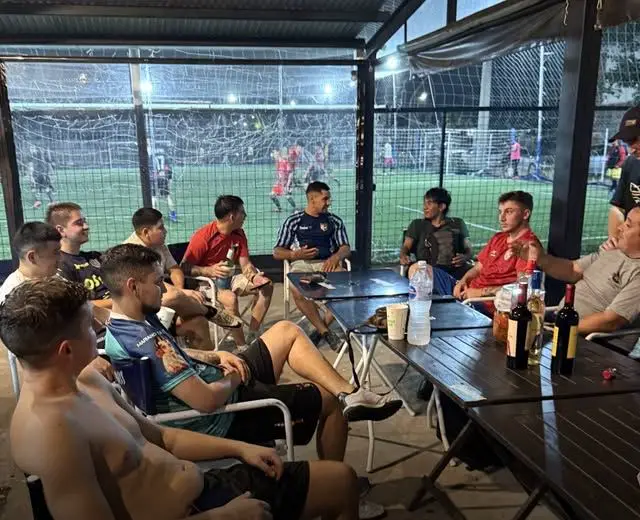 Jugadores en las mesas del deck techado, con la cancha iluminada de fondo